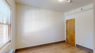 This single-floor unit presents a clean, functional layout finished with warm light hardwood flooring and crisp white walls throughout, creating a bright and consistent aesthetic from room to room. The main living area is an open, versatile space anchored by a wood-framed entry door and ceiling-mounted lighting fixtures. A wall-mounted radiator sits beneath a window dressed with horizontal blinds, allowing natural light to filter in while offering views of the surrounding greenery outside. The simplicity of the room's design makes it highly adaptable for various living configurations. A dedicated closet provides practical built-in storage, featuring a wood hanging rod and shelving ideal for organization. The bathroom is thoughtfully appointed with light hardwood-style flooring that continues the unit's cohesive design language. It includes a wall-mounted sink, toilet, a framed mirror, and a separate walk-in shower stall with grab bars, suggesting accessibility-conscious design. A frosted window allows natural light into the bathroom while maintaining privacy. Dark wood baseboards add a subtle contrast throughout the unit, lending a touch of warmth to the otherwise neutral palette. Overall, the space offers a straightforward, well-maintained environment suited for efficient, everyday living.
