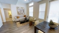 This well-appointed single-floor residence offers a thoughtfully designed open-concept layout that seamlessly connects the living and dining areas. The living space is anchored by large windows dressed with white blinds that flood the room with natural light, creating an airy and welcoming atmosphere. Durable wood-look flooring in a warm grey tone extends throughout, providing both style and practicality. The living area features elevated ceilings with a subtle tray detail, adding architectural interest and enhancing the sense of vertical space. A large abstract canvas in gold and grey tones serves as a striking focal point on the accent wall, complementing the neutral color palette of warm whites and beige. The space is well-suited for both relaxing and entertaining, with a generous seating arrangement and an adjacent dining area highlighted by a dark wood table. Recessed lighting and ceiling-mounted fixtures provide consistent illumination throughout. The overall ambiance is modern yet inviting, striking a balance between clean, contemporary design and warm, livable comfort — making it an ideal space for everyday living.