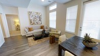 This well-appointed single-floor residence offers a thoughtfully designed open-concept layout that seamlessly connects the living and dining areas. The living space is anchored by large windows dressed with white blinds that flood the room with natural light, creating an airy and welcoming atmosphere. Durable wood-look flooring in a warm grey tone extends throughout, providing both style and practicality. The living area features elevated ceilings with a subtle tray detail, adding architectural interest and enhancing the sense of vertical space. A large abstract canvas in gold and grey tones serves as a striking focal point on the accent wall, complementing the neutral color palette of warm whites and beige. The space is well-suited for both relaxing and entertaining, with a generous seating arrangement and an adjacent dining area highlighted by a dark wood table. Recessed lighting and ceiling-mounted fixtures provide consistent illumination throughout. The overall ambiance is modern yet inviting, striking a balance between clean, contemporary design and warm, livable comfort — making it an ideal space for everyday living.