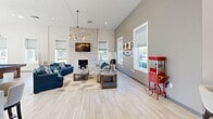 Located at 901 W Little Creek Rd in Norfolk, Virginia, this single-floor community clubhouse offers a thoughtfully designed, open-plan layout with a welcoming and modern aesthetic. Light hardwood-style flooring flows throughout, complementing the soaring ceilings and abundant natural light streaming through large, Roman shade-dressed windows.

The expansive main area serves multiple functions, anchored by a striking stacked-stone fireplace with a wall-mounted television, framed by plush blue sofas and casual seating — an inviting lounge ideal for relaxing. A pool table adds a recreational dimension to the space, alongside a classic popcorn cart that lends a playful touch.

The kitchenette is a standout feature, centered around a large quartz-topped island with upholstered seating. Elegant gold geometric pendant lights hang above, while the backsplash wall features a bold striped tile pattern. Stainless steel appliances, ample cabinetry, and a full-size refrigerator complete the well-equipped prep area.

A glass-walled private conference room offers a dedicated workspace with a central table, task seating, computers, and a sculptural pendant light. A cozy seating nook with a round glass table basks in natural light from corner windows with outdoor views. The space proudly displays a "We Support Our Troops" banner, reflecting its community-oriented character.