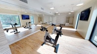This single-floor fitness studio is designed specifically for indoor cycling and group exercise, offering a clean, contemporary environment conducive to high-energy workouts. The space features light hardwood flooring throughout, which adds warmth while remaining practical for fitness use. A full wall of floor-to-ceiling mirrors lines one side of the room, visually expanding the space and allowing participants to monitor their form during sessions. Multiple professional-grade stationary cycling bikes are arranged across the studio floor, providing ample room between each unit for comfortable use. Pendant lighting fixtures hang from the white ceiling at regular intervals, casting an even, well-distributed light across the room. A mounted flat-screen television is positioned on the mirrored wall, visible from all cycling positions. The far wall features a textured stone or tile accent panel, adding architectural interest and contrast to the otherwise minimalist palette. Built-in shelving and cabinetry along this accent wall offer organized storage for equipment and accessories. Natural light enters from a window near the television wall, enhancing the bright and welcoming atmosphere. The overall design is clean and functional, promoting focus and motivation.