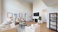 Located at The Villas at Setauket in East Setauket, NY, this two-story villa offers a thoughtfully designed layout with high-end finishes across both floors. The main living area immediately impresses with soaring double-height ceilings, warm hardwood flooring, and a gas fireplace as a central focal point. A staircase with a wood handrail connects the two levels, adding architectural character to the open layout.

The dining area is bathed in natural light from multiple windows framed with white blinds, and is anchored by a striking industrial-style orb chandelier above a substantial wood dining table. The kitchen features white shaker cabinetry extending to the ceiling, crisp quartz countertops, and stainless steel appliances including a gas range, over-the-range microwave, and a French door refrigerator.

The bedrooms are generously proportioned, with the primary suite offering hardwood flooring and ample natural light, while a secondary bedroom features plush carpeting and bright window access. The bathroom is finished with marble-patterned tile flooring, a white vanity with a dark countertop, and a tub-shower combination with elegant marble-style surround tile. An attached garage with an automatic opener completes this well-appointed home.