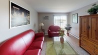 This single-floor residence offers a thoughtfully organized layout that accommodates a variety of living needs. The living room serves as the central gathering space, featuring plush red leather seating, a glass-top coffee table, and a wall-mounted air conditioning unit, with a large window fitted with vertical blinds that allow natural light to filter in. Adjacent to the living area, the dining room provides a cozy setting with a round wooden table and four chairs, accented by framed botanical artwork on the walls and wood-style flooring that continues into the kitchen. The kitchen is equipped with warm oak cabinetry, granite-style countertops, a double stainless steel sink, and a dishwasher, offering ample storage and prep space. The primary bedroom features a classic wooden bed frame, a matching dresser with mirror, sliding door access, and soft carpeting. Additional bedrooms are furnished with wooden bunk beds and natural light from windows with blinds, one also including a mirrored vanity desk. The bathroom presents wood-look vinyl flooring, a granite-topped vanity with oak cabinetry, and a tub-shower combination. Throughout the residence, neutral wall tones and warm wood furnishings create a consistent, welcoming atmosphere.