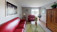 This single-floor residence offers a thoughtfully organized layout that accommodates a variety of living needs. The living room serves as the central gathering space, featuring plush red leather seating, a glass-top coffee table, and a wall-mounted air conditioning unit, with a large window fitted with vertical blinds that allow natural light to filter in. Adjacent to the living area, the dining room provides a cozy setting with a round wooden table and four chairs, accented by framed botanical artwork on the walls and wood-style flooring that continues into the kitchen. The kitchen is equipped with warm oak cabinetry, granite-style countertops, a double stainless steel sink, and a dishwasher, offering ample storage and prep space. The primary bedroom features a classic wooden bed frame, a matching dresser with mirror, sliding door access, and soft carpeting. Additional bedrooms are furnished with wooden bunk beds and natural light from windows with blinds, one also including a mirrored vanity desk. The bathroom presents wood-look vinyl flooring, a granite-topped vanity with oak cabinetry, and a tub-shower combination. Throughout the residence, neutral wall tones and warm wood furnishings create a consistent, welcoming atmosphere.
