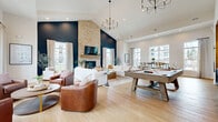 The Kingsley Park Apartments clubhouse at 5151 Kingsley Park Dr in Columbus, Ohio, is a thoughtfully designed community amenity space that combines comfort, function, and sophisticated style across a single expansive floor. The welcoming entryway sets an immediate tone with its bold deep-blue walls, herringbone hardwood flooring, curated wall art, and glass-paneled office suite visible from the entrance corridor. The main lounge area is an architectural standout, featuring soaring vaulted ceilings, a dramatic floor-to-ceiling natural stone fireplace flanked by a navy accent wall, and an array of large windows that fill the room with abundant natural light and views of the surrounding community. Comfortable seating arrangements, warm leather accent chairs, and a statement chandelier contribute to the inviting ambiance. A billiards table and a built-in bar area with navy cabinetry, exposed brick backsplash, open shelving, and pendant lighting provide excellent social and entertainment opportunities, complemented by bar-height seating for casual gatherings. The dedicated office space offers a quiet, professional setting with glass walls and functional furnishings. The fitness center is generously equipped with cardio machines, strength training equipment, free weights, and rubber sport flooring beneath vaulted ceilings, with large windows overlooking the outdoor pool area.