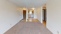 This single-floor residential unit offers a practical and comfortable layout designed for everyday living. The spacious living room is finished with wall-to-wall carpeting and features a textured ceiling, neutral walls, and a sliding glass door that opens to an outdoor balcony area, allowing natural light to fill the room and providing pleasant views of the surrounding greenery.

The open layout connects the living area seamlessly to the kitchen, which is equipped with warm-toned natural wood cabinetry, a dishwasher, a refrigerator, and a range. A window above the sink brings in additional natural light, while tile flooring adds a clean, durable surface underfoot. A baseboard heating unit ensures year-round comfort throughout the space.

A hardwood-floored entryway and hallway connect the main living spaces to the private areas of the unit, including the bedroom and bathroom. The full bathroom features a clean, white tub-shower combination with glass enclosure, a toilet, and a vanity with a dark wood cabinet base and an integrated sink, complemented by a mirrored medicine cabinet above.

The 3D dollhouse view reveals a compact yet efficient floor plan that maximizes usable space, with clearly defined zones for living, cooking, and sleeping, making this a well-organized and functional residential unit.