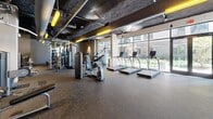 Located at 101 Nerem St NW in Atlanta, GA, this well-appointed fitness center offers a comprehensive workout environment across a single, open floor. The space is thoughtfully divided into distinct exercise zones, catering to a wide range of fitness activities.

The cardio area is a standout feature, lined with floor-to-ceiling windows that flood the space with natural light and provide views of the surrounding outdoor courtyard. This section is equipped with a row of treadmills, elliptical trainers, stair climbers, and recumbent bikes from the TRUE Fitness brand, each outfitted with individual display screens.

A dedicated strength and free weight area features a full dumbbell rack, flat and adjustable benches, and cable machines. Full-length mirrors amplify the sense of space and allow for proper form checks during workouts. A separate enclosed room with glass walls houses additional cable and multi-functional strength machines, offering a more focused training environment.

The industrial-style ceiling, left intentionally exposed to reveal ductwork, piping, and dark-painted surfaces, is accented by striking yellow pendant lighting that adds a modern, energetic character to the space. Durable rubber flooring throughout ensures safety and functionality. The fitness center also includes a dedicated cycling studio with stationary bikes and media screens, completing this versatile and well-equipped facility.