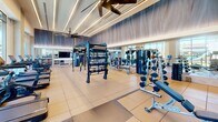 Located at 18750 Delaware St in Huntington Beach, California, this fitness center offers a generously sized, single-floor exercise space designed to accommodate a comprehensive range of workout needs. The facility features soaring ceilings accented by sleek linear lighting and large ceiling fans, creating an airy and energizing environment throughout.

Floor-to-ceiling windows line one side of the room, flooding the space with abundant natural light and providing open views of the surrounding Huntington Beach streetscape. The interior is finished with large-format tile flooring complemented by rubber mat zones for functional training areas.

The cardio section is equipped with multiple treadmills, stationary bikes, and rowing machines, all positioned to take advantage of the natural light and outdoor views. The strength training area is equally well-appointed, featuring a multi-functional cable and functional trainer rig, a full dumbbell rack, barbells, weight benches, and a variety of selectorized machines. Medicine balls and additional accessories round out the functional fitness offerings.

Full-length mirrors are strategically placed throughout the space, enhancing the sense of depth while supporting proper form during workouts. A flat-screen television is mounted for entertainment. The facility's contemporary design, with its textured wall paneling and modern finishes, delivers a polished and professional fitness environment.
