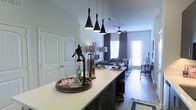 This single-floor space showcases a thoughtfully designed open-concept layout that blends the kitchen, living, and dining areas into one cohesive and functional environment. The kitchen features a generous island with a light-toned countertop and dark cabinetry, offering ample prep and seating space. Three striking black geometric pendant lights hang above the island, adding a bold design statement that anchors the kitchen zone. The flooring throughout is a warm hardwood tone that unifies the open space and adds a sense of continuity. The living area is bathed in natural light, courtesy of large windows dressed with layered curtains and blinds, creating a bright yet comfortable atmosphere. A ceiling fan overhead provides both practicality and a subtle design touch. A glass-paneled door at the far end of the living area invites additional light and provides access to the exterior. The dining area, visible to the right, complements the overall aesthetic with coordinating furnishings. The neutral color palette of soft whites and grays throughout the space creates a clean, contemporary feel, while carefully chosen décor accents add warmth and personality. The seamless open floor plan makes the space feel expansive and well-connected, ideal for both everyday living and entertaining.