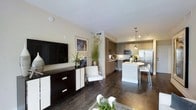 This single-floor space showcases a thoughtfully designed open-concept layout that connects the living and kitchen areas in a seamless, functional flow. Rich, dark hardwood flooring extends throughout, lending warmth and continuity to the overall design.

The living area features neutral-toned walls that create a clean, versatile backdrop, complemented by a wall-mounted flat-screen television positioned above a two-toned dark wood and white media console. Framed artwork and lush greenery add visual interest and a welcoming ambiance to the space.

Transitioning directly into the kitchen, the design showcases two-toned cabinetry combining warm wood uppers with grey lower cabinets, paired with light countertops. A central island with a white base provides additional prep space and casual seating via modern bar stools. Stainless steel appliances, including a French door refrigerator and built-in microwave, contribute to the kitchen's contemporary character. Pendant lighting suspended above the island offers both task illumination and a decorative focal point.

Overall, the space presents a cohesive modern aesthetic, balancing functional design with refined finishes throughout.