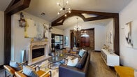 This Houston lobby presents a striking blend of rustic and contemporary design elements across its single-level layout. The interior immediately draws attention upward to soaring vaulted ceilings adorned with substantial dark wood beams, including a dramatic arched beam detail that serves as a central architectural focal point. Sputnik-style chandeliers provide ambient and accent lighting throughout, complementing wall-mounted lantern sconces that add a classic touch.

The entry foyer features rich slate tile flooring transitioning to warm wood-look flooring in the main lounge area. Exposed brick walls with arched passageways lend an old-world character, framing a pair of solid wood double entry doors fitted with an arched transom window. Frosted glass sliding barn doors offer connectivity between distinct zones, providing both visual interest and functional separation.

The lounge area is anchored by an ornate carved stone fireplace topped with a large-scale abstract artwork. Seating is thoughtfully arranged with a dark leather sofa, patterned accent chairs in amber and teal tones, and geometric-print area rugs. Additional abstract wall art, decorative consoles, and curated accessories complete the refined aesthetic. Windows along one wall allow natural light to filter in, enhancing the lobby's welcoming ambiance.