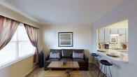 This one-bedroom, one-bathroom unit is thoughtfully designed to maximize its compact footprint across a single floor. The living area serves as the heart of the space, featuring warm hardwood-style flooring, neutral beige walls, and a smooth white ceiling that enhances the sense of openness. A window dressed with rich, patterned curtains in deep burgundy and brown tones admits natural light, brightening the room. The living space comfortably accommodates a full-sized sofa and coffee table, creating an inviting area for relaxation.

A standout architectural detail is the pass-through opening that connects the living room to the kitchen, fostering an open, communicative flow between the two spaces. The kitchen features white cabinetry and a countertop bar ledge with seating for bar stools, offering a casual dining option. This pass-through design adds visual depth and functionality without sacrificing the defined layout of each area.

Overall, the unit presents a practical and comfortable living environment with a warm, neutral palette and efficient use of space throughout.
