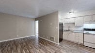 This efficient one-bedroom, one-bathroom unit is thoughtfully laid out across a single floor, offering a practical and comfortable living environment. The open-concept living and kitchen area forms the heart of the space, finished with wood-look plank flooring in a warm, neutral tone that extends throughout. The kitchen is positioned at one end, featuring white cabinetry, a stainless steel refrigerator, a white electric range, a single-basin sink with granite-look countertops, and a window that brings in natural light. A textured ceiling and neutral taupe-toned walls give the living area a clean, understated aesthetic. The bedroom is a well-proportioned private retreat equipped with a wall-mounted air conditioning unit, a window with blinds, and a double closet with built-in rods, offering generous storage. The bathroom includes a vanity with a granite-look countertop, a mirrored medicine cabinet, and a separate water closet, all finished with the same durable wood-look flooring. A wall-mounted HVAC unit and floor vent provide climate control throughout the unit. The overall layout is efficient and cohesive, making practical use of every square foot.