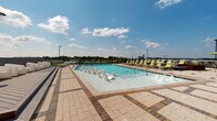 This rooftop amenity deck offers a well-appointed outdoor retreat centered around a generously sized swimming pool with crystal-clear water. The pool features in-water lounge chairs positioned in the shallow end, allowing residents to relax while partially submerged, alongside traditional pool entry steps with stainless steel railings. The surrounding terrace is finished in a patterned mix of light and warm-toned pavers, creating a polished, contemporary aesthetic across the expansive deck surface.

Rows of yellow-green sling-style chaise lounges line the perimeter of the pool area, providing ample sunbathing space. A raised wooden deck platform with modern white lounge chairs offers an additional seating vignette, ideal for socializing or relaxing away from the water. Shaded dining areas with umbrellas and outdoor tables are situated near the adjacent clubhouse structure, which features floor-to-ceiling windows and glass doors that connect interior amenity spaces to the outdoor deck.

A glass-paneled railing borders portions of the terrace, preserving unobstructed panoramic views of the surrounding tree canopy and urban skyline. Colorful potted flowers add seasonal accents throughout. The overall ambiance is modern, open, and resort-inspired, benefiting from abundant natural light and sweeping open-air views.