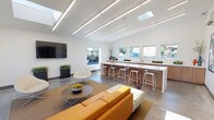 This contemporary single-floor space is designed with a clean, open-plan layout that seamlessly integrates a lounge area and a full kitchen bar. The interior is defined by crisp white walls and polished concrete floors, creating a bright, minimalist aesthetic throughout.

The lounge area is anchored by a vibrant orange sofa paired with a round coffee table and a modern swivel chair, complemented by a wall-mounted flat-screen television and colorful abstract artwork. Large glass doors along one wall flood the space with natural light and provide direct access to an outdoor patio, blurring the boundary between interior and exterior.

The kitchen bar is a standout feature, stretching nearly the full length of the room with a long white quartz countertop, warm wood cabinetry beneath, and a row of sleek white metal barstools. A secondary preparation counter lines the back wall, equipped with a sink, dishwasher, and additional storage, with a series of horizontal windows above offering views to the outside.

Overhead, parallel linear LED lighting runs across the ceiling alongside strategically placed skylights, ensuring the entire space is evenly and warmly illuminated at all times. A split-system air conditioner is mounted on the wall, providing climate control. The overall ambiance is modern, welcoming, and highly functional, making this space well-suited for social gatherings or community use.
