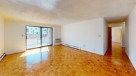 Located at 75 Morton Village Dr in Boston's Mattapan neighborhood, this single-floor residence offers a practical and well-organized layout throughout. The living areas are unified by warm, polished parquet wood flooring in a classic block pattern that runs consistently through the main rooms, reflecting natural light and adding visual warmth to the space. Baseboard heating runs along the perimeter walls, and a wall-mounted air conditioning unit provides climate control year-round.

The living room is generously proportioned, featuring a large sliding glass door that opens to a balcony with outdoor views, flooding the space with natural light. An additional windowed room provides further brightness and serves well as a bedroom or secondary living area.

The galley-style kitchen is efficiently designed, featuring warm-toned wood cabinetry both above and below white countertops, with under-cabinet lighting over the range. White appliances include a refrigerator, electric range, sink, and dishwasher, all set atop neutral tile flooring.

The bathroom is a notable standout, showcasing a full tub-and-shower combination clad entirely in retro aqua-blue ceramic tile, lending a distinctive mid-century character to the space. The textured ceilings are consistent throughout the unit, completing the overall aesthetic.