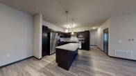This single-floor unit showcases a contemporary design centered around a striking open-concept kitchen and living area. The kitchen is a standout feature, outfitted with rich dark cabinetry, granite countertops, and a full suite of stainless steel appliances, including a refrigerator, range, microwave, and dishwasher. A decorative diamond-pattern wine rack adds a distinctive detail above the sink area. The spacious central island with a granite surface provides additional prep space and serves as a natural divider between the kitchen and the generous open living area, which features light wood-style plank flooring throughout.

The living space is bright and versatile, with neutral walls and recessed lighting complementing the pendant lights above the island. Bedrooms are well-proportioned, with wood-style flooring, natural light from window treatments, and dark-trimmed doors that echo the kitchen's design palette.

The unit includes two bathrooms, one of which is ADA-accessible, featuring grab bars, an open under-sink vanity with granite countertops, and a tub/shower combination. The second bathroom mirrors the same finishes with dark cabinetry and a tub/shower unit. The cohesive design elements throughout create a polished, modern aesthetic.