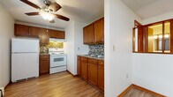 This single-floor unit offers a practical and cohesive layout connecting its key living spaces. The kitchen is a standout area, showcasing warm medium-toned wood cabinetry with brushed nickel hardware, granite-look countertops, and a mosaic tile backsplash that adds visual texture. White appliances, including a range, oven, and refrigerator, are neatly integrated alongside a stainless steel sink. A ceiling fan with a light fixture overhead provides both ventilation and ambient lighting, while a textured ceiling adds subtle character throughout. Wood-look laminate flooring flows seamlessly through the kitchen and into the open living area, which benefits from multiple windows that bring in natural light and views of surrounding greenery. A wall-mounted air conditioning unit and baseboard heating provide year-round climate control. The living space is open and versatile, offering ample room for various furniture arrangements. A separate bedroom features neutral carpet and clean white walls, providing a quiet, comfortable retreat. The bathroom is finished with white square wall and floor tile, a full soaking tub with a shower, an over-toilet cabinet for additional storage, and wood-look flooring that ties back to the rest of the unit.