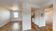 This single-floor unit offers a practical and straightforward layout connecting a living area, kitchen, bedroom, and bathroom. The living area is finished with wood-style hard flooring throughout, neutral-toned walls, and a textured ceiling. A window fitted with horizontal blinds allows natural light to fill the space, while a flush-mount ceiling light fixture provides overhead illumination. A partial wall separates the living area from the kitchen corridor, adding a degree of visual definition between the two spaces.

The galley-style kitchen is efficiently arranged with white cabinetry lining both sides, offering ample upper and lower storage. Laminate countertops provide functional workspace, and the kitchen is equipped with black appliances including a refrigerator, electric range with microwave above, and a dishwasher. A track lighting fixture overhead brightens the workspace.

The bedroom features soft carpeting, a ceiling fan with light kit, and a window that draws in natural light. The bathroom includes a vanity with a speckled countertop, a tub and shower combination enclosed by sliding glass doors, and textured walls. Wood-style flooring continues into the bathroom, maintaining visual consistency throughout the unit.