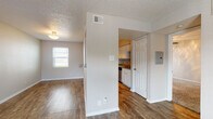 This single-floor unit offers a practical and straightforward layout connecting a living area, kitchen, bedroom, and bathroom. The living area is finished with wood-style hard flooring throughout, neutral-toned walls, and a textured ceiling. A window fitted with horizontal blinds allows natural light to fill the space, while a flush-mount ceiling light fixture provides overhead illumination. A partial wall separates the living area from the kitchen corridor, adding a degree of visual definition between the two spaces.

The galley-style kitchen is efficiently arranged with white cabinetry lining both sides, offering ample upper and lower storage. Laminate countertops provide functional workspace, and the kitchen is equipped with black appliances including a refrigerator, electric range with microwave above, and a dishwasher. A track lighting fixture overhead brightens the workspace.

The bedroom features soft carpeting, a ceiling fan with light kit, and a window that draws in natural light. The bathroom includes a vanity with a speckled countertop, a tub and shower combination enclosed by sliding glass doors, and textured walls. Wood-style flooring continues into the bathroom, maintaining visual consistency throughout the unit.