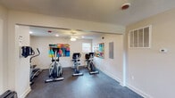 This fitness center in Baton Rouge, Louisiana, is a single-story facility designed to provide a functional and visually engaging workout environment. The layout features two connected exercise areas that flow seamlessly from an entry corridor into a broader open workout space, offering ample room for cardio training and general exercise.

The flooring throughout the facility consists of durable speckled rubber, well-suited for high-traffic fitness use. White walls and recessed lighting create a clean, bright atmosphere, while ceiling fans in both sections of the space promote air circulation during workouts. Windows positioned along the walls allow natural light to filter in, enhancing the overall openness of the facility.

Cardio equipment is a central feature of the space, including Matrix treadmills, elliptical trainers, and stationary bikes arranged to maximize usability. An open section of the larger room provides additional floor space for stretching or bodyweight exercises.

A notable design element is the collection of large, colorful abstract artworks mounted throughout the space, adding a lively and dynamic aesthetic to the otherwise neutral interior. A dedicated entry area with a branded welcome mat, exit signage, and a glass-panel door completes the space, contributing to its organized and accessible layout.