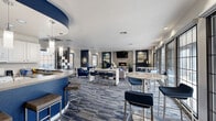 This well-appointed community lounge and kitchen space is designed with a modern, cohesive aesthetic centered around a striking navy blue and white color palette. The open-plan layout accommodates multiple functional zones that flow seamlessly into one another, all unified by patterned blue-toned carpeting throughout.

At the heart of the kitchen area, a sweeping curved bar island finished in bold navy serves as a dramatic focal point, framed by a matching arched ceiling detail above. White cabinetry, pendant lighting, and full appliances complete this fully equipped kitchen, which offers ample bar seating for casual gatherings.

The living area is generously furnished with a mix of plush sofas, tufted navy accent chairs, and marble-top bistro tables, offering versatile seating arrangements for both relaxed lounging and social interaction. A stone-clad fireplace wall anchors one end of the room, topped by a wall-mounted flat-screen television and flanked by curated artwork and decorative shelving.

A dedicated lounge nook features a sleek open shelving unit and large grid-pane windows that flood the space with natural light and provide views of the surrounding outdoor area. An entry corridor with glass door access connects the interior directly to an outdoor patio, further extending the space's versatility. Throughout, thoughtful décor including greenery, framed artwork, and decorative accessories adds warmth to the modern design.