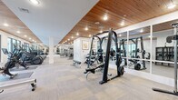 This fitness center offers a thoughtfully designed workout environment that balances functional utility with refined architectural detail. The space is organized across a single floor, providing generous room for a comprehensive range of exercise equipment and activities.

The ceiling is a standout architectural feature, finished with warm-toned natural wood planking and recessed lighting that creates an inviting, upscale atmosphere. White structural columns are positioned throughout, complementing the clean, neutral color palette of the walls and the light-toned modular flooring below.

Large floor-to-ceiling mirrors run along key wall sections, visually expanding the space and reflecting both the equipment and natural light entering through exterior-facing windows. The strength training area is well-equipped with Smith machines, multi-station cable systems, free weights, and adjustable benches by Precor and similar professional-grade brands. A dedicated cardio zone accommodates multiple treadmills, rowing machines, and a stationary cycle.

Additional storage is provided through built-in white cabinetry and an open shelving unit housing medicine balls and accessories. The entrance corridor leads smoothly into the main floor, completing a well-organized, premium fitness facility.