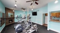 This single-floor fitness center is designed to support a comprehensive workout experience in a welcoming and well-maintained environment. The space is immediately distinguished by its vibrant aqua-colored walls, white trim, and dark rubber flooring, creating a clean and energizing atmosphere throughout.

The main cardio area is generously sized and houses a variety of professional-grade equipment, including Precor ellipticals, treadmills, a WaterRower rowing machine, and upright stationary bikes. Dual ceiling fans provide comfortable airflow, while pendant lighting adds a warm, inviting touch. A wall-mounted flat-screen television is positioned for easy viewing during workouts. A full-wall mirror runs along one side, enhancing the sense of space and allowing users to monitor their form.

A charming brick fireplace with a mantle adds a distinctive architectural element, lending character to what might otherwise be a purely utilitarian space.

The strength training room offers a multi-functional cable machine, a tiered dumbbell rack with a full range of free weights, a workout bench, and additional mirror panels along the wall. Natural light enters through slatted windows, keeping the area bright and airy.

Adjacent to the main workout areas is a functional kitchenette, featuring warm wood cabinetry, a gas range, ample countertop space, a microwave, a water cooler, and a refrigerator — ideal for post-workout refreshment and convenience.