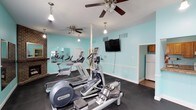 This single-floor fitness center is designed to support a comprehensive workout experience in a welcoming and well-maintained environment. The space is immediately distinguished by its vibrant aqua-colored walls, white trim, and dark rubber flooring, creating a clean and energizing atmosphere throughout.

The main cardio area is generously sized and houses a variety of professional-grade equipment, including Precor ellipticals, treadmills, a WaterRower rowing machine, and upright stationary bikes. Dual ceiling fans provide comfortable airflow, while pendant lighting adds a warm, inviting touch. A wall-mounted flat-screen television is positioned for easy viewing during workouts. A full-wall mirror runs along one side, enhancing the sense of space and allowing users to monitor their form.

A charming brick fireplace with a mantle adds a distinctive architectural element, lending character to what might otherwise be a purely utilitarian space.

The strength training room offers a multi-functional cable machine, a tiered dumbbell rack with a full range of free weights, a workout bench, and additional mirror panels along the wall. Natural light enters through slatted windows, keeping the area bright and airy.

Adjacent to the main workout areas is a functional kitchenette, featuring warm wood cabinetry, a gas range, ample countertop space, a microwave, a water cooler, and a refrigerator — ideal for post-workout refreshment and convenience.