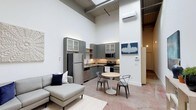 Located at 1200 S 2nd St in Wilmington, NC, this single-floor unit showcases a striking open-concept design defined by dramatically high ceilings that lend an airy, loft-like atmosphere. Exposed structural elements overhead, including industrial-style beams and ductwork, complement the crisp white walls, creating a compelling contrast between raw and refined aesthetics. The polished concrete-style flooring flows continuously throughout, reinforcing the industrial character of the space.

The kitchen is efficiently laid out along one wall, featuring gray-toned cabinetry with glass-front upper cabinets, stainless steel appliances—including a full-size refrigerator, microwave, and range—and a functional work counter. A compact dining area sits adjacent to the kitchen, furnished with a round pedestal table and mixed metal chairs, making it an ideal spot for casual meals.

The living area provides generous room for lounging, anchored by a sectional sofa and area rug. A wall-mounted mini-split HVAC unit ensures year-round climate control. Skylights and elevated windows draw in natural light, enhancing the overall sense of openness. Decorative artwork adds a curated, welcoming touch.