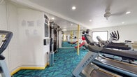 This fitness center is a comprehensively outfitted exercise facility designed to support a full range of workout routines. The space is laid out on a single level and features a bold, eye-catching teal geometric carpet that energizes the room and sets a vibrant tone throughout. Recessed lighting illuminates the interior evenly, complemented by a ceiling fan and natural light streaming in through windows along one wall.

The cardio section is well-appointed with multiple Matrix treadmills arranged in a row, an elliptical trainer, and an upright stationary bike, offering ample options for aerobic exercise. A multi-station cable strength machine anchors the strength training area, while a red adjustable weight bench and an assortment of stability balls — in red and black — provide versatile options for functional and free-weight training.

Full-length mirrors line one wall, amplifying the sense of space and allowing users to monitor their form during workouts. Yellow accent columns add a striking pop of color that ties into the yellow baseboard trim, creating a cohesive and lively aesthetic. Decorative wall art, including a geode-inspired piece and graphic prints, adds personality to the space. Informational signage is posted near the entrance for user reference. The overall ambiance is bright, clean, and motivating.