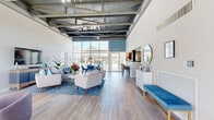 Located along Oak Trail S in Clearwater, Florida, this single-story community clubhouse delivers a bright, welcoming atmosphere enhanced by thoughtful modern design. The main gathering area commands immediate attention with its dramatic exposed beam ceiling, soaring height, and a bold blue accent wall that adds a vibrant coastal character. Floor-to-ceiling windows and glass entry doors flood the interior with abundant natural light while framing views of the outdoor pool area and lush Florida landscaping beyond.

The open main hall features light wood-tone flooring throughout, creating a seamless flow between functional zones. A seating area with contemporary mid-century styled furniture, accented by blue upholstery and patterned area rugs, provides a comfortable gathering space. A dedicated reception desk near the entry offers a welcoming point of contact, accompanied by stylish purple accent chairs and warm wood finishes.

Adjacent to the main hall, a fully equipped kitchen showcases dark cabinetry, granite countertops, stainless steel appliances, and ample workspace, ideal for community events. A secondary lounge area provides additional seating alongside a media console, while a glass-walled fitness room with cardio equipment is conveniently accessible nearby. Together, these spaces create a cohesive, well-appointed community facility.