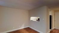 This single-floor space presents a clean, minimalist layout with a notable architectural detail: a pass-through window cut into the wall connecting the main living area to the kitchen. This practical design element allows for visual connectivity between the two spaces while maintaining a degree of separation. The living area features warm-toned hardwood flooring that adds character and depth to the room, complemented by smooth, neutral beige walls that create a bright and versatile backdrop. White baseboards provide a crisp contrast against both the walls and flooring, contributing to a polished finish throughout. The kitchen, partially visible through the pass-through window, includes a modern faucet and counter space. The ceiling is smooth and uniform, enhancing the overall sense of openness in the room. Electrical outlets are thoughtfully positioned along the walls, offering practical functionality. The neutral color palette and straightforward layout make the space highly adaptable for various furnishing styles and uses. Overall, the space feels tidy, well-maintained, and move-in ready, with its pass-through window serving as a functional and visually interesting centerpiece.