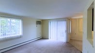 This single-floor residential space offers a straightforward and functional layout designed for comfortable everyday living. The main living area is well-lit, benefiting from multiple windows that allow natural light to fill the room, complemented by a set of sliding glass doors dressed with vertical blinds that provide both privacy and access to the outdoors. The flooring throughout is wall-to-wall carpeting in a neutral tone, contributing to a warm and quiet atmosphere. Climate comfort is addressed through a combination of a wall-mounted air conditioning unit and baseboard heating running along the perimeter of the room, ensuring year-round temperature control. The walls are finished in a soft, neutral cream tone that enhances the brightness of the space and creates a clean, understated aesthetic. The open layout of the living area provides flexibility for a variety of furniture arrangements, making it an adaptable environment. Overall, the space presents a simple, practical interior with functional amenities suited to residential use.