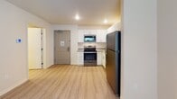 Located at 9844–9930 59th Ave N in Plymouth, Minnesota, this single-floor unit offers a clean, modern interior with consistent light wood-tone flooring throughout. The kitchen is a functional highlight, outfitted with white cabinetry, granite-style countertops, and a full suite of stainless steel appliances including a refrigerator, electric range, over-the-range microwave, and dishwasher. Recessed lighting and ample cabinet storage make this an efficient and practical cooking space. The kitchen opens directly into the living area, creating an easy flow between spaces. The living area is bright and open, with a window dressed in blinds that allows natural light to filter in. The bedroom features neutral-toned walls with a cool gray accent wall, a window with curtains for privacy, and generous floor space. The bathroom is well-appointed with white cabinetry, granite-style countertops, a full tub-shower combination, and a towel rack, all finished with coordinating hardware. The unit's overall design is contemporary and neutral, with textured ceilings, clean white walls, and consistent flooring that ties each space together cohesively.