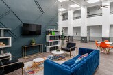 Located at 300 W 2nd St in Santa Ana, California, this single-floor space makes a striking impression with its dramatic double-height ceilings and an abundance of natural light streaming through multiple skylights overhead. The open floor plan is anchored by a bold, slate-blue geometric accent wall that serves as a focal point, adorned with a wall-mounted television and flanked by white-and-gold shelving units. Plush navy-blue velvet sofas, gold-framed accent chairs, and a vibrant multicolored area rug create an inviting lounge area that balances style with comfort. Ceiling fans are suspended from the towering ceiling, enhancing both aesthetics and airflow throughout the space. Structural columns clad in gray bases punctuate the open interior, adding architectural character while defining distinct zones. A dining area furnished with white tables and vibrant orange chairs provides a casual setting for gatherings, while high-top tables with modern seating offer additional flexible workspace. Wood-look tile flooring runs throughout, tying the space together seamlessly. The overall ambiance is contemporary and energetic, with a thoughtful mix of bold color, modern furnishings, and architectural grandeur that makes this a dynamic and functional community environment.