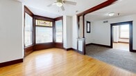 Located at 220 Iowa Ave in Muscatine, Iowa, this 2-bedroom, 1-bathroom unit blends historic architectural charm with functional living. The space is defined by rich, dark-stained hardwood floors that run throughout, complemented by bold wood baseboards, detailed window casings, and ornate decorative columns that frame the transition between rooms — all hallmarks of classic early 20th-century craftsmanship.

The combined living area is bright and open, highlighted by a striking bay window adorned with colorful stained glass transom panels that cast warm, decorative light into the room. A ceiling fan adds comfort, and the open flow between areas creates a spacious, airy feel. The kitchen features white cabinetry, granite-look countertops, white appliances including a refrigerator, dishwasher, and electric range, and grey wood-look flooring that transitions cleanly from the hardwood in the living area.

Both bedrooms offer hardwood floors, multiple windows for natural light, and classic wood-trimmed doors. The primary bedroom is generously sized with a corner bay window and stained glass accent, offering views of the surrounding area. The bathroom includes a white vanity with cabinet storage, a well-lit mirror, wood-look flooring, and accessible grab bars. Throughout the unit, the consistent use of dark trim against white walls creates a timeless, character-rich aesthetic.