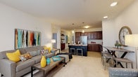This single-floor residence is designed with an open-concept layout that seamlessly connects the living area and kitchen, creating a spacious and functional flow. The living room is bright and inviting, benefiting from large windows that allow generous natural light to fill the space. A tiled fireplace serves as a focal point, complemented by plush carpeting underfoot. A glass-paneled door provides additional light and access to the exterior.

The kitchen is a standout feature, outfitted with rich dark-stained cabinetry extending to the ceiling, light granite countertops, and a suite of stainless steel appliances including a refrigerator, range, and over-the-range microwave. A contrasting dark island with granite counters and bar seating anchors the space, illuminated by pendant lighting above. Light wood-style flooring runs throughout the kitchen, transitioning to carpet in the adjoining living areas.

The bedroom is comfortably scaled, featuring carpeted flooring, a large window with soft drapery, and ample space for a full dresser ensemble. The bathroom features a warm wood vanity with white countertops, a large mirror, and clean, neutral finishes throughout.
