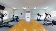 Located at 101 Chandler Rd in Greer, South Carolina, this single-floor community amenity facility offers residents a well-rounded set of shared spaces designed for convenience and leisure. The fitness center serves as a centerpiece of the building, featuring gleaming hardwood flooring down a wide central corridor flanked by carpeted exercise zones. Equipment including a treadmill, elliptical, and stationary bike is positioned to maximize open floor space, while large windows along the exterior wall allow natural light to fill the area. The light gray walls and bright overhead lighting contribute to a clean, welcoming atmosphere throughout. A dedicated screening room provides an immersive entertainment experience, furnished with rows of plush, dark reclining theater-style chairs equipped with cup holders and surrounded by wall-mounted surround sound speakers. Soft sconce lighting and patterned carpeting complete the cinematic ambiance of this intimate viewing space. The laundry room is bright and functional, with a large window overlooking the surrounding community, in-unit washer and dryer units, and ample space for comfortable use. Together, these amenities reflect a thoughtfully designed facility that supports the everyday lifestyle needs of its community.