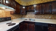 Located at 3704 Main St in Mineral Ridge, Ohio, this single-floor unit offers a straightforward, functional layout across several distinct areas. The kitchen is one of the more characterful spaces, outfitted with rich, dark-stained wood cabinetry that provides ample upper and lower storage. A textured brick-patterned wall treatment adds visual interest, complementing the white countertops, double basin sink, electric range, and dishwasher. The vinyl tile flooring in the kitchen provides a durable, easy-to-clean surface.

The living areas and bedrooms feature wall-to-wall carpeting in a neutral tan tone, contributing a warm, understated feel throughout. Multiple rooms are served by baseboard heating units running along the perimeter walls, ensuring consistent warmth. Each carpeted room includes windows that bring in natural light and offer outdoor views of the surrounding area, with one room also featuring a wall-mounted air conditioning unit for cooling.

The bathroom is equipped with a full tub-and-shower combination, surrounded by beige tile surround panels. A dark wood vanity with a marbled countertop and vinyl flooring complete the space, offering a cohesive, practical design. Overall, the unit presents a clean, no-frills interior with consistent finishes throughout.