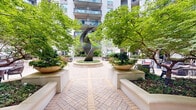 Located at 6205 Carnegie Blvd in the SouthPark area of Charlotte, NC, this beautifully designed outdoor courtyard serves as the communal centerpiece of a multi-story residential building. The space is defined by its expansive herringbone-patterned brick paver plaza, which guides residents through thoughtfully arranged seating areas, including woven lounge chairs with red cushions, round dining tables with coordinating chairs, and curved built-in benches with a clean, contemporary finish.

Lush, mature landscaping is a defining feature throughout the courtyard, with ornamental trees providing natural canopy shade and vibrant flowering plants nestled in raised garden beds. The verdant greenery creates a sense of enclosure and tranquility, offering a private retreat within the urban setting. A central fountain serves as a focal point visible from multiple vantage points within the space.

The surrounding multi-story building facade features large sliding glass doors and windows at the ground level, opening directly onto the courtyard and reinforcing a seamless indoor-outdoor connection. Private balconies with iron railings line the upper floors, overlooking the garden below. Subtle landscape lighting and sculptural art elements add visual interest, completing an outdoor environment designed for relaxation and community engagement.