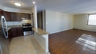 This single-floor space offers an open-concept design that seamlessly connects the kitchen and living area, creating a cohesive and functional layout. The kitchen is equipped with stainless steel gas range and dark espresso cabinetry, providing a strong contrast against the light-toned granite countertops. A peninsula with matching granite extends from the kitchen, serving as a natural divider between the kitchen and living space while offering additional prep or dining surface. The kitchen features tile flooring, while the adjoining living area transitions to rich, dark hardwood-style flooring, adding warmth and visual definition to the space. Neutral-toned walls throughout keep the interior feeling bright and versatile. A range hood above the stove ensures practical ventilation. The living area is generously proportioned, offering ample room for flexible furniture arrangements. A doorway visible in the background hints at additional rooms beyond, suggesting a well-connected floor plan. Recessed lighting in the kitchen, combined with the open layout, contributes to an overall sense of brightness. The space presents a clean, move-in-ready aesthetic with a balance of practicality and modern design sensibility.