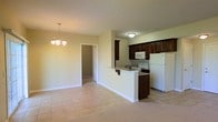 This single-floor space showcases an open-concept design that seamlessly connects the living, dining, and kitchen areas into one cohesive layout. The kitchen is equipped with dark espresso-toned cabinetry that provides a rich contrast against the light-colored walls and neutral flooring. White appliances, including a refrigerator, range, and over-the-range microwave, are built into the kitchen space. A peninsula counter with a breakfast bar overhang extends into the main living area, offering additional seating and workspace while maintaining visual flow between zones. The dining area is defined by a hanging chandelier with a classic multi-bulb design, adding a subtle decorative focal point to the space. Flooring transitions from tile in the kitchen and main living zone to carpet in the adjoining areas, including what appears to be a hallway leading to additional rooms. The walls are painted in a warm, neutral tone throughout, contributing to a cohesive and understated ambiance. White baseboards and standard ceiling heights complete the clean, functional aesthetic of this straightforward, practical layout suitable for everyday living.