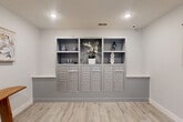 Located at 1301 Rudel Drive in Tomball, Texas, this hallway serves as a functional and thoughtfully designed mailroom space. The single-floor layout is efficiently organized, offering a welcoming atmosphere despite its compact footprint.

The centerpiece of the space is a full-wall built-in mailbox unit finished in a sleek gray tone. It features multiple individual mail compartments, larger parcel lockers, open display shelving above, and flanking marble-topped counter surfaces — providing both utility and visual appeal. Decorative accents, including ceramic vases and a white orchid arrangement, add a polished, curated touch to the mailbox wall.

The entry side of the hallway features a glass-paneled exterior door that allows natural light to filter in, complemented by recessed ceiling lighting throughout. A live-edge wooden bench positioned beneath an abstract framed artwork adds a warm, artistic element, while a lush fiddle-leaf fig tree in a matte black planter introduces a natural, organic accent. A world map wall hanging adds an additional layer of character to the space.

Underfoot, light wood-look plank flooring runs throughout, enhancing the bright, airy feel of the space. The walls are finished in a soft neutral gray, contributing to the clean, contemporary aesthetic. Overall, this hallway balances practical function with a refined, modern sensibility.