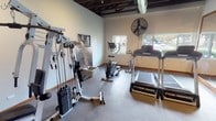 This single-floor fitness space offers a practical and well-equipped environment for a variety of workout routines. The room features an industrial-style aesthetic, with an exposed ceiling showcasing dark-painted structural beams, visible ductwork, and track lighting that provides focused illumination throughout the space. The flooring consists of durable interlocking rubber or foam tiles, well-suited for exercise use and easy maintenance.

The fitness area is outfitted with multiple cardio machines, including two treadmills and an elliptical trainer, positioned to take advantage of the natural light streaming in through a row of windows along one wall. These windows offer views of the surrounding outdoor environment, contributing to an open, airy atmosphere. A large pedestal fan is mounted on the wall to assist with ventilation and airflow during workouts.

A large framed mirror spans one wall, visually expanding the space and allowing users to monitor their form during exercise. An accent wall features exposed brick detailing, adding a textural contrast to the otherwise smooth, light-painted walls. The space also includes a multi-function cable weight machine for strength training, along with an exercise reference poster. Additional open floor space provides room for stretching or bodyweight exercises, making this a versatile and functional fitness facility.