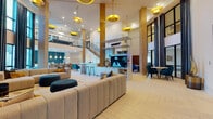 This two-story community clubhouse in Lakewood Ranch, Florida, is designed with a bold contemporary aesthetic that blends vibrant color accents with modern architectural details. The main level opens through sleek black-framed glass double doors into a grand, double-height atrium anchored by soaring ceilings and warm wood-clad columns. Striking gold saucer pendant lights hang overhead, casting a warm glow across herringbone-patterned light tile flooring that flows throughout the main gathering areas.

The open-plan first floor is organized into distinct social zones. A central media unit with a flat-screen TV and built-in shelving serves as a visual divider between a bar-height counter with gold-accented stools and an intimate dining cluster with teal velvet chairs. Tufted gold booth seating adds a lounge-bar feel, while floor-to-ceiling windows framed by dramatic navy curtains flood the space with natural light and offer views of palm-lined surroundings. A small beverage station with teal herringbone tile backsplash provides added functionality near the entrance corridor.

A secondary lounge area features vibrant teal carpeting, a tufted charcoal sofa, a scalloped velvet accent chair, and framed coastal artwork. The showpiece of the space is a dramatic full-wall mural in teal, copper, and gold abstract tones anchoring a curved sectional lounge. A staircase with black metal railings connects to the second-floor mezzanine level, visible from the main atrium below.