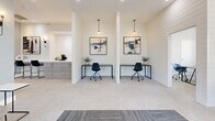 Located at 316 Columbia Dr in Carrollton, GA, this single-floor co-working space offers a clean, modern aesthetic designed to support focused productivity. The space is divided into two distinct office areas, each thoughtfully appointed with sleek, minimalist furnishings and consistent design details throughout.

The primary workspace features striking white shiplap accent walls adorned with framed artwork, pendant lighting, and individual desks paired with ergonomic rolling chairs. Soft, neutral carpeting runs underfoot, contributing to a calm and professional atmosphere. A small refreshment station with cabinetry is conveniently positioned nearby, adding a practical amenity for occupants.

The secondary office area is surrounded by large, multi-pane windows that flood the room with natural light and offer views of the surrounding community. Additional workstations provide flexible seating arrangements suitable for individual or small-group use. Recessed ceiling lighting supplements the natural light, ensuring a well-illuminated environment at all times.

The entry area features a mix of patterned and solid carpet tile, a glass entry door, and ceiling fan, creating a welcoming and functional transition into the workspace. A private office or consultation room with a large window and a long wooden table rounds out the offering, ideal for focused meetings or private work sessions. Together, these spaces deliver a polished, professional environment well-suited for remote work or collaborative use.