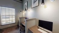 Located at 5501 Old Montgomery Hwy in Tuscaloosa, Alabama, this single-floor business center offers a thoughtfully designed environment built to support both focused work and informal meetings. The space is divided into two distinct functional zones, each with its own carefully considered aesthetic.

The workstation area features multiple built-in desks with decorative column dividers, providing semi-private individual work alcoves. Each station is outfitted with task-appropriate lighting via sleek black and gold adjustable wall sconces, and desktop computers are available for use. A central window with horizontal blinds allows natural light to filter into the room, while abstract framed artwork adorns the soft gray-green walls, lending a refined, professional atmosphere. Patterned carpet underfoot adds warmth and texture throughout.

Adjacent to the workstation zone is a cozy lounge seating area anchored by a round table and striped upholstered chairs. This nook is distinguished by a dramatic wall installation of convex circular mirrors in varying sizes, creating a visually dynamic focal point. A bold color-wheel artwork adds a vibrant accent, while a pendant light overhead provides warm, ambient illumination. Crown molding details throughout the space reinforce a polished, upscale design sensibility.