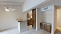 This single-floor space showcases an open and functional layout that seamlessly connects the living area to the kitchen. The living space features plush carpeting in a neutral tone, complemented by crisp white walls that create a bright and airy atmosphere. Exposed ceiling beams painted white add a subtle architectural character to the room, lending visual interest overhead.

A built-in wooden cabinet unit along one wall provides convenient storage and display space, crafted in a warm medium-toned wood that adds a natural element to the interior. A peninsula-style counter with a practical overhang extends from a dividing wall, creating a natural pass-through between the living and kitchen areas while offering additional counter or bar seating potential.

The kitchen, visible through a wide pass-through opening framed by a rustic wood-accented door surround, is equipped with matching medium-toned wood cabinetry, a range, and tile flooring. Upper and lower cabinets provide ample storage, and a range hood is positioned above the cooking area. A ceiling vent ensures proper climate control throughout the space. The cohesive use of warm wood tones and neutral whites throughout creates a welcoming and understated ambiance.