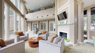 This sophisticated community lounge space is defined by its dramatic double-height ceilings and an abundance of natural light streaming through expansive floor-to-ceiling windows. The windows offer direct views of a resort-style outdoor pool and lush surrounding greenery, creating a seamless connection between indoor comfort and outdoor amenity. The main lounge area is anchored by a stately white-mantled fireplace, above which a wall-mounted flat-screen television is positioned. Decorative framed accents adorn the mantel, adding a refined, curated touch. Comfortable gray upholstered seating, accented with warm amber throw pillows, is arranged around a rich cherry wood circular coffee table, all set atop a patterned area rug with an elegant interlocking motif. Upper-level clerestory windows line the walls, further enhancing the open and airy atmosphere of the space. A separate bar-height seating area is visible toward the rear, outfitted with bistro-style stools and a high table, ideal for casual gatherings. French door sets lead to additional rooms and outdoor access points. The overall aesthetic blends classic architectural detailing—including crown molding and columnar accents—with contemporary furnishings, resulting in a welcoming and polished communal environment.